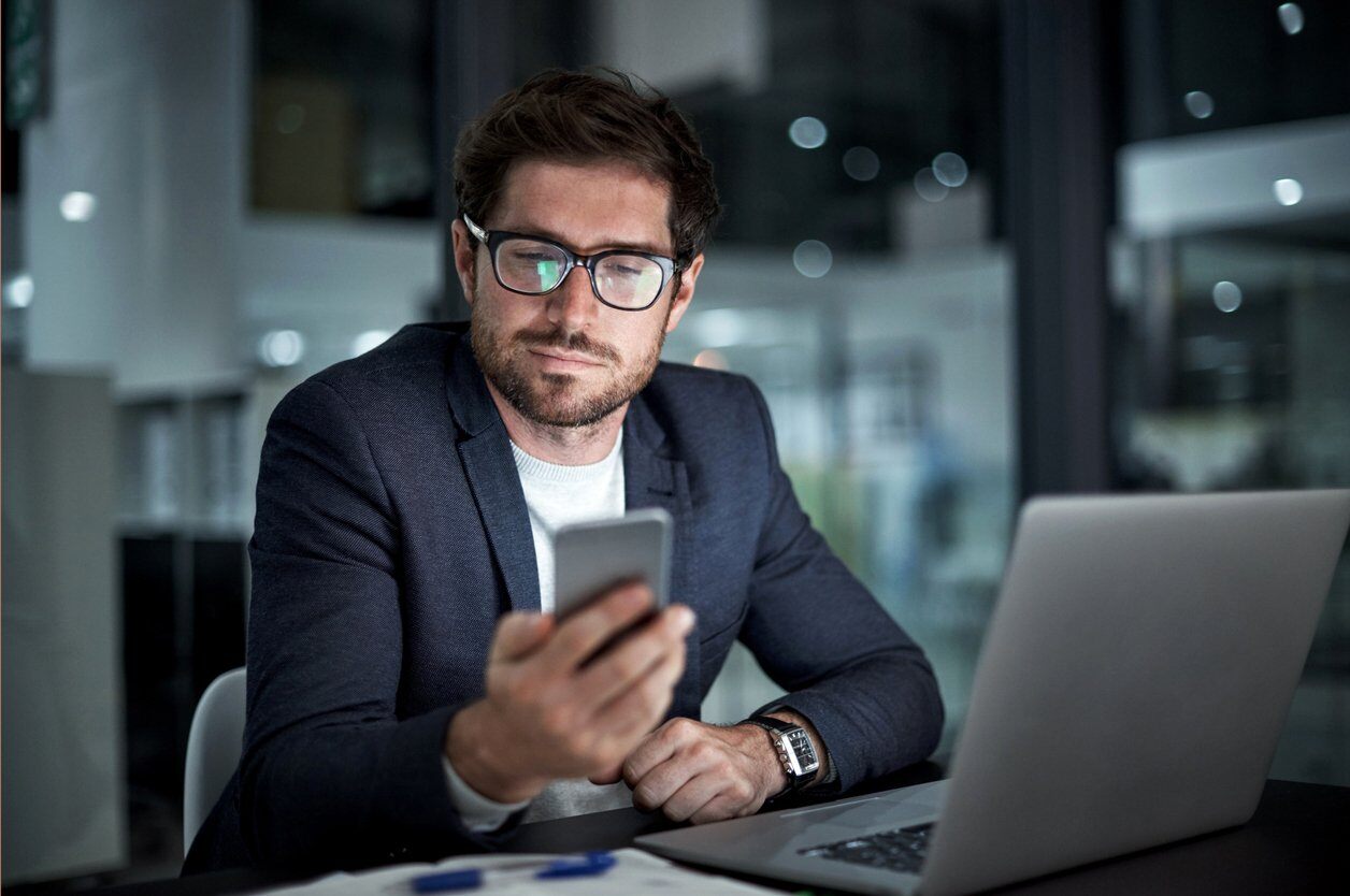 Guy sitting infront of his laptop and looking at his phone